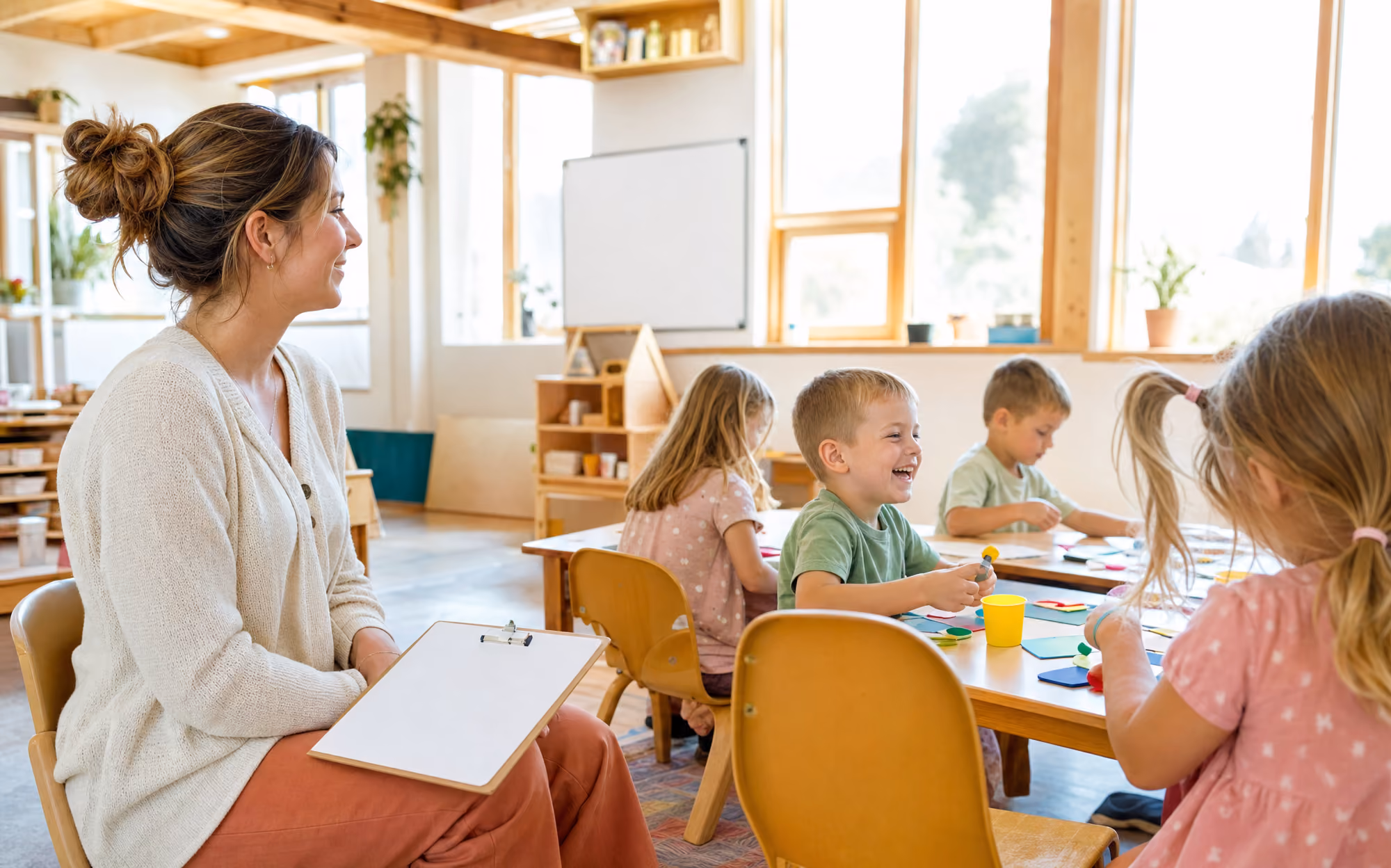 An adult sitting at the side of a primary-school classroom taking observation notes while children work at their desks.